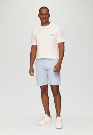 Light beige t-shirt with short sleeves and blue text, paired with light blue shorts, and white sneakers. Smooth fabric, casual fit.
