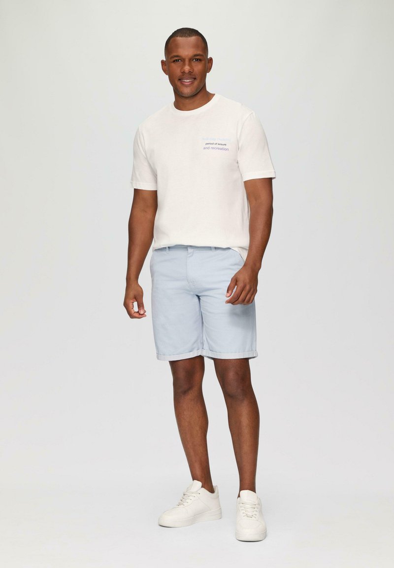 Light beige t-shirt with short sleeves and blue text, paired with light blue shorts, and white sneakers. Smooth fabric, casual fit.