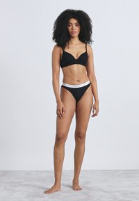 Μαύρο σουτιέν και ταιριαστό σλιπ μπικίνι, με λευκή ζώνη μέσης με λογότυπο, λεία υφή και απλό σχέδιο. Το μοντέλο στέκεται σε ανοιχτό γκρι επιφάνεια.