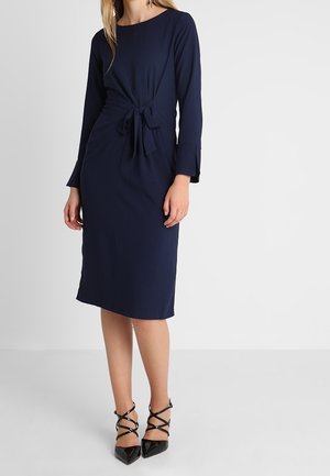 Robe bleu marine longueur genou avec manches longues, détail noué à l'avant et légère fente latérale, portée avec des escarpins noirs à brides et bouts pointus.