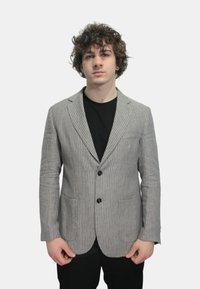 Blazer a righe in tessuto grigio e bianco, con un solo bottone, revers classici e tasche frontali. Indossato sopra una camicia nera.