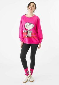 Fuchsia Sweatshirt mit einem Snoopy-Print, kombiniert mit schwarzen Leggings und leuchtend pinkfarbenen Socken mit Charakterdesign. Weiße Sneakers sind enthalten.
