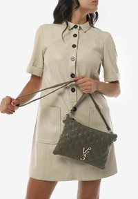 Borsa a tracolla in pelle verde oliva con texture e motivo geometrico, dotata di accessori in oro. Il modello indossa un vestito beige con colletto.