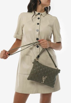 Borsa a tracolla in pelle verde oliva con texture e motivo geometrico, dotata di accessori in oro. Il modello indossa un vestito beige con colletto.