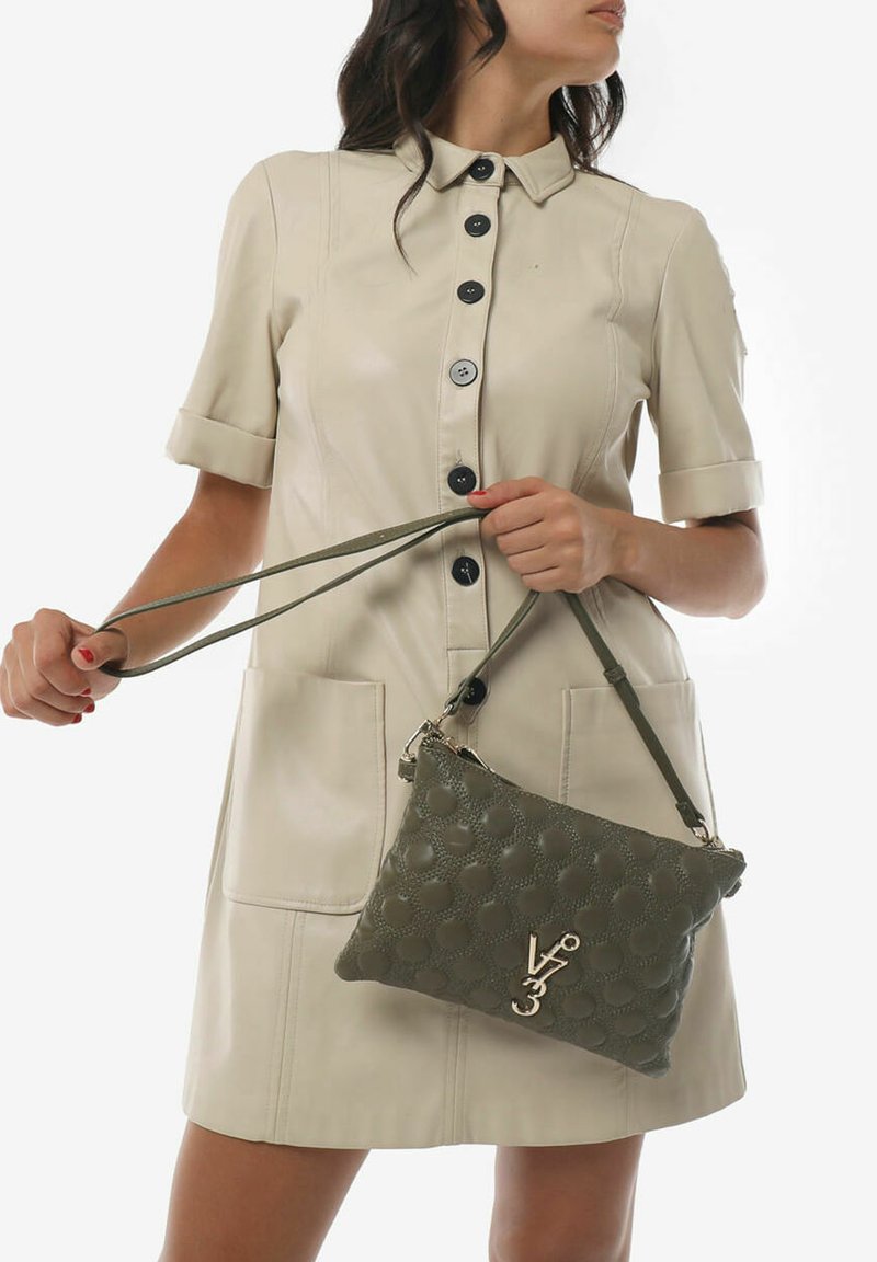 Borsa a tracolla in pelle verde oliva con texture e motivo geometrico, dotata di accessori in oro. Il modello indossa un vestito beige con colletto.