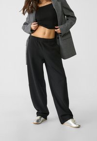 Zwarte wijde broek met een elastische tailleband, gecombineerd met een zwarte geribde crop top en een grijze blazer, aangevuld met zilveren sneakers.