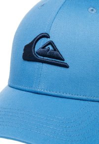 Blue cap with a structured design, featuring a navy embroidered logo and textured fabric. The peak is flat with visible stitching details.