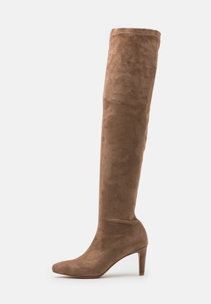 Wysokie damskie brązowe zamszowe kozaki z wąskim noskiem i średniej wysokości szpilką, sięgające do kolan, na jednolitym białym tle.
