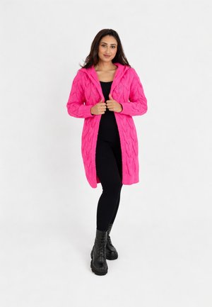 Kobieta stojąca w jaskrawo różowym kapturowym kardiganie z warkoczowym splotem, czarnym topie i legginsach oraz czarnych butach sznurowanych na białym tle.