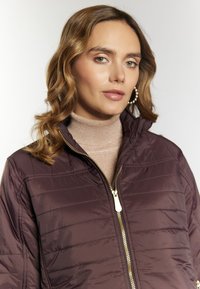 Giacca bordeaux trapuntata con colletto alto, cerniera dorata e cuciture orizzontali. Indossata sopra un maglione a collo alto chiaro e luccicante.