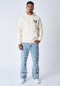 Sudadera crema con un diseño de mariposa negra en el pecho, combinada con unos jeans desgastados de color azul claro que presentan roturas horizontales y zapatillas blancas.
