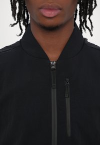 Black bomber jacket with a minimal design, featuring a standing collar and two zippered pockets at the front. Smooth texture, lightweight fabric.
