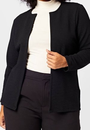 Kvinde iført en sort ribbet åben cardigan over en hvid højhalset top og sorte bukser, der holder cardigan med den ene hånd.