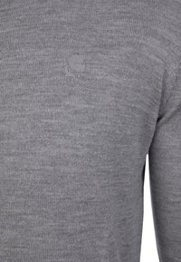 Grå stickad tröja med ribbad struktur, rund halsringning och subtil logotypdetalj. Tyget ser mjukt och figurnära ut.