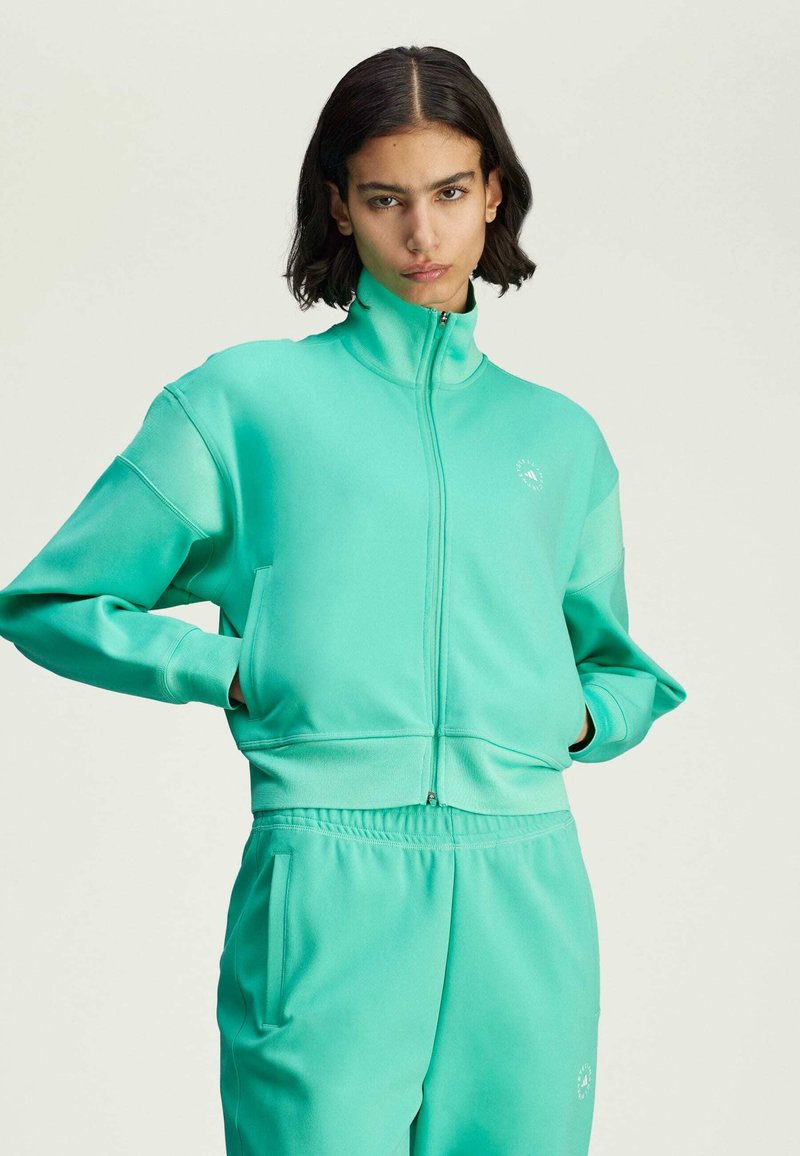 Giacca zip-up turchese chiaro realizzata in tessuto liscio, con colletto alto e due tasche laterali. Dettaglio del logo semplice sul petto.