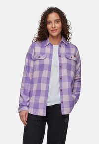Camicia di flanella a scacchi viola e rosa chiaro con due taschini sul petto e bottoni marroni, indossata sopra una maglietta bianca, abbinata a pantaloni neri.