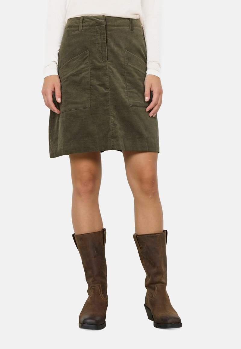 Femme portant une jupe droite en velours côtelé vert olive avec poches à l'avant, un haut blanc à manches longues et des bottes en cuir marron mi-mollet, debout.