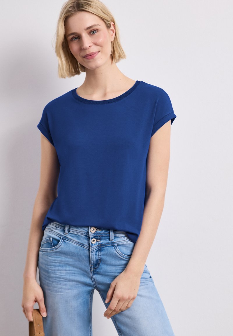 Femme aux cheveux blonds courts portant un t-shirt bleu roi à manches courtes et un jean bleu clair, tenant une ceinture marron dans la main gauche.