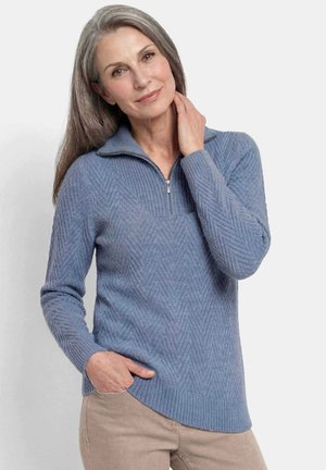 Femme mûre aux cheveux gris porte un pull bleu en maille avec fermeture éclair et un pantalon beige, posant avec une main dans la poche et l'autre touchant son cou.
