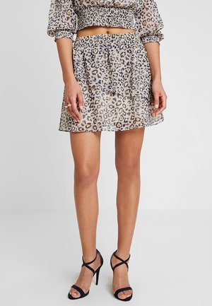 Femme portant une jupe courte à imprimé léopard avec un top court assorti et des sandales noires à talons hauts, debout devant un arrière-plan uni.