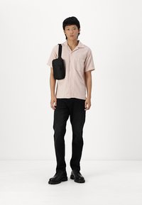 Camisa de manga corta a rayas en rosa, pantalones negros y zapatos negros. Bolso cruzado en negro. Fondo liso.