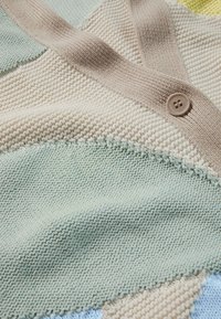 Neulevaate, joka esittelee kermansävyä, mintunvihreää ja pehmeää keltaista, tekstuurilla varustetuilla kuvioilla ja ruskealla nappiyksityiskohdalla.