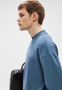 Μπλε φούτερ με ριμπ λαιμόκοψη, απαλό ύφασμα, σχέδιο με χαμηλωμένους ώμους. Μαύρη τσάντα ακουμπάει στο πλάι. Απλό φόντο.