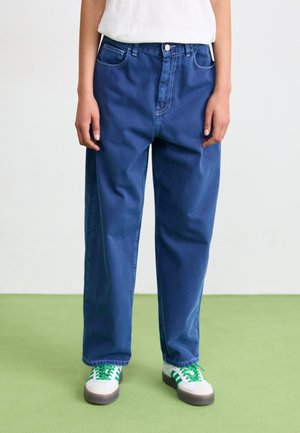 Osoba ubrana w luźne niebieskie dżinsy i białe trampki z zielonymi paskami, stojąca na zielonej podłodze na tle jednolitej białej ściany.