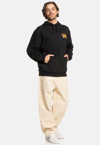 Sudadera negra con bolsillo frontal, capucha con cordón y gráfico colorido en el pecho. Combinada con pantalones holgados crema y zapatos blancos.
