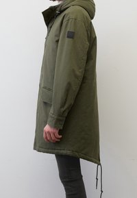 Olivgrön parkas med huva, som har en framzip, knäppte manschetter och en patch på vänster ärm. Tyget har en mjuk struktur.