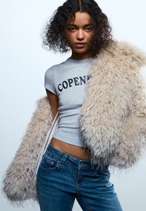 Jauna sieviete ar cirtainiem matiem valkā pelēku T-kreklu ar uzrakstu "Kopenhāgena", zilas džinsas un tur pār pleciem bēšas pūkainas virsjakas.