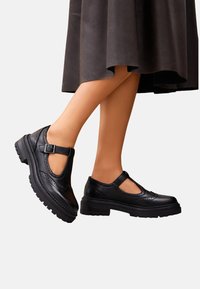 Des chaussures T-strap en cuir noir présentent un bout arrondi, des détails perforés et une semelle en caoutchouc épaisse. Boucle réglable pour un ajustement parfait.