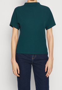 Tyrkysový top s krátkým rukávem a vysokým výstřihem, vyrobený z hladkého materiálu. Nosit s tmavými džíny, které mají rovný střih.