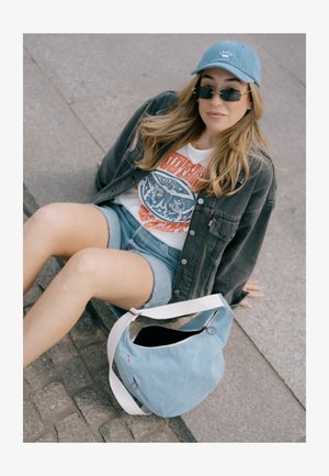 Bolsa de ombro em denim azul claro com uma alça branca, apresentando um fecho de correr e detalhes de ferragens circulares. Ambiente urbano casual.