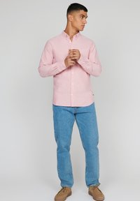 Lichtroze overhemd met lange mouwen, gecombineerd met blauwe denim jeans en bruine schoenen. Het overhemd heeft een klassieke kraag en een subtiele textuur.