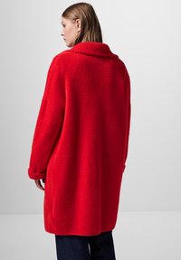 Röd, överdimensionerad kappa i texturerat tyg, med en bred krage och uppvikta ärmar, som sträcker sig till mitten av låret. Enkel, enfärgad design.