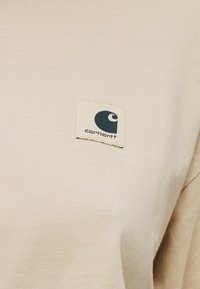 Μπεζ πουκάμισο Carhartt με μικρό τετράγωνο λογότυπο στο στήθος, που απεικονίζει ένα μαύρο μισοφέγγαρο και το όνομα της μάρκας.