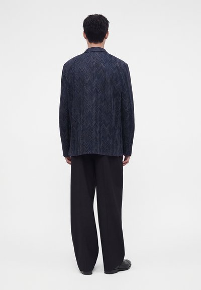 Homme debout de dos portant une veste bleu foncé à motif chevrons, un pantalon noir à jambes larges et des chaussures noires sur fond blanc.