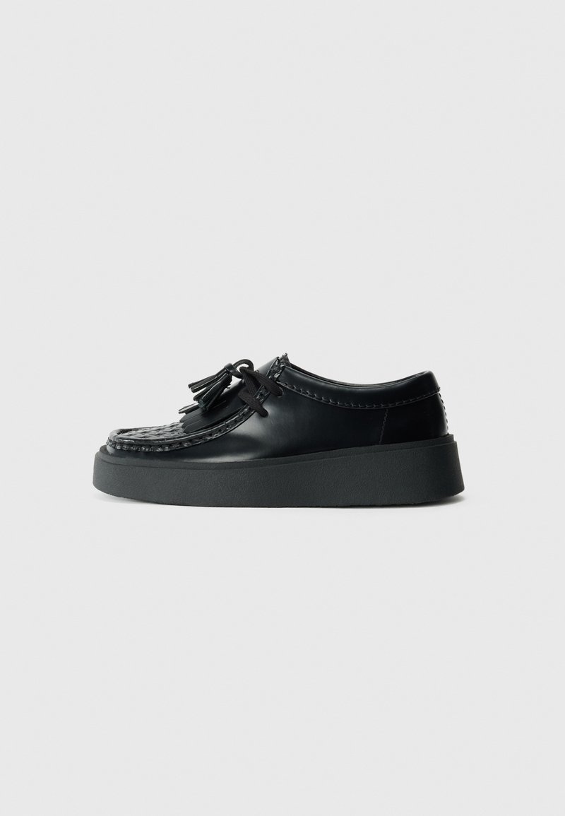 Black leather shoes with a woven toe detail, black laces, and a thick black rubber sole, featuring tassels as a notable accent.