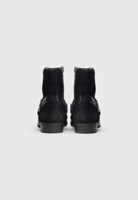 Černé semišové kotníkové boty se zipy na zadní straně, nízkými podpätky a ozdobnými pásky. Hladká textura s jemnými detaily na páscích.