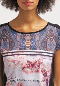 Pestrobarevná krátká halenka s květinovými a vzorovanými potisky, s černým výstřihem a vodorovnými pruhy na lemu.