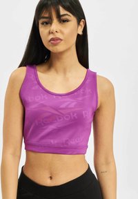 Fialový športový crop top s vzorom loga Reebok, s okrúhlym výstrihom a hladkým, elastickým materiálom. Pod ním sú čierne nohavice s vysokým pásom.