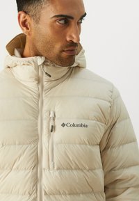 Beige puffer jacket with hood, featuring a front zipper and Columbia logo on the chest. Quilted design with soft texture.