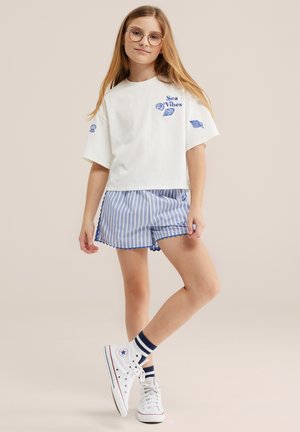 Noor tüdruk pikkade juustega, prillidega, valge "Sea Vibes" T-särgiga, siniste triibuliste lühikeste pükste, triibuliste sokkidega ja valgete tennistega, seisab lõdvestunult.