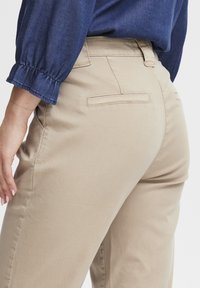 Beige chinos met zijzakken en twee achterzakken, met een gladde textuur. Een blauwe denim blouse met pofmouwen is gedeeltelijk zichtbaar.