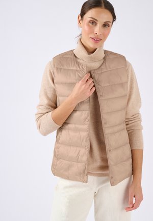Mujer con chaleco acolchado beige sobre un suéter de cuello alto beige y pantalones blancos, abrochando el chaleco con la mano derecha.