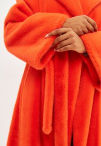 Rochie din blană artificială portocalie cu mansete largi, o curea și o textură moale. Prezintă un aspect pufos și o nuanță vibrantă pentru un interes vizual.