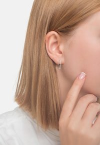 Silver geometriska örhängen med en elegant vertikal design, visade på en modells öra, med kort blont hår och en ljusfärgad topp.