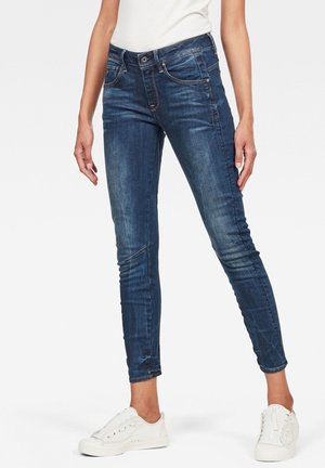 Jean slim bleu foncé porté avec des baskets blanches et un haut blanc sans manches, visible de la taille jusqu’aux pieds sur un fond uni.