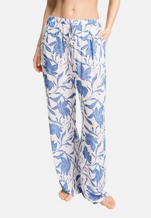 Vrouw draagt witte losse broek met blauwe bloemenprint en elastische tailleband met trekkoord, staat blootsvoets met één hand in de zak.